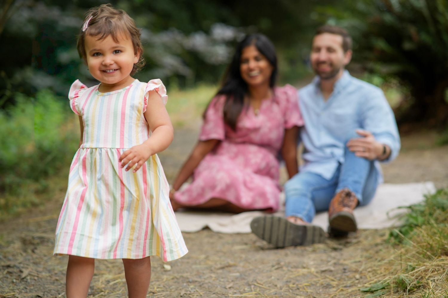 Young girl smiling and walking toward the camera while her parents sit together on a blanket outdoors.