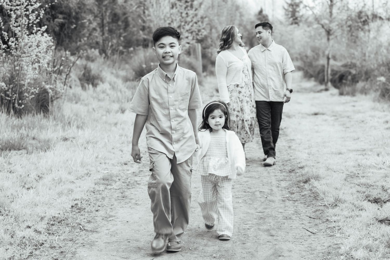 Black and white portrait of siblings walking hand in hand while their parents follow behind.
