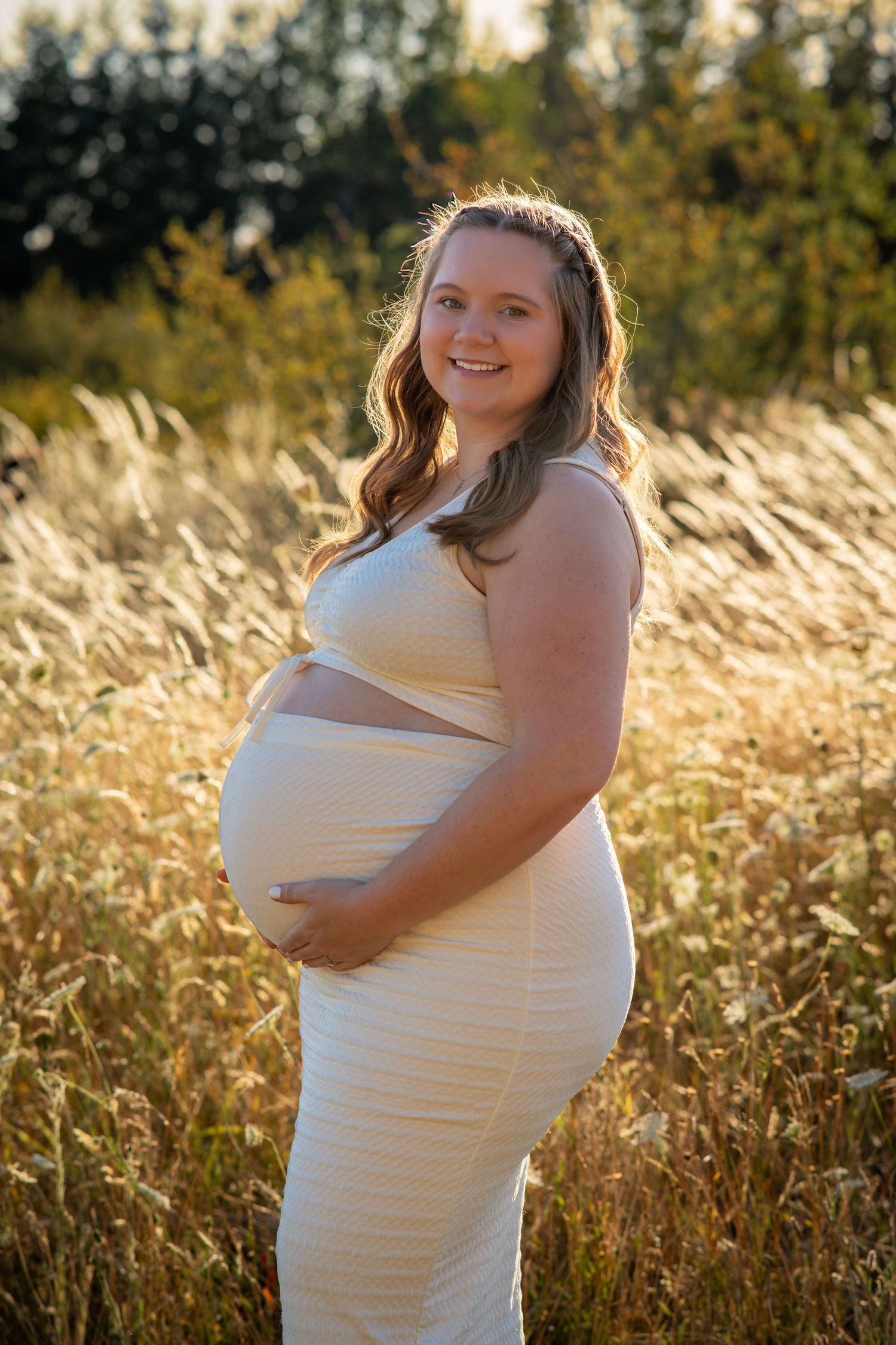 Pregnant woman smiling and cradling her baby bump in a golden sunlit field.