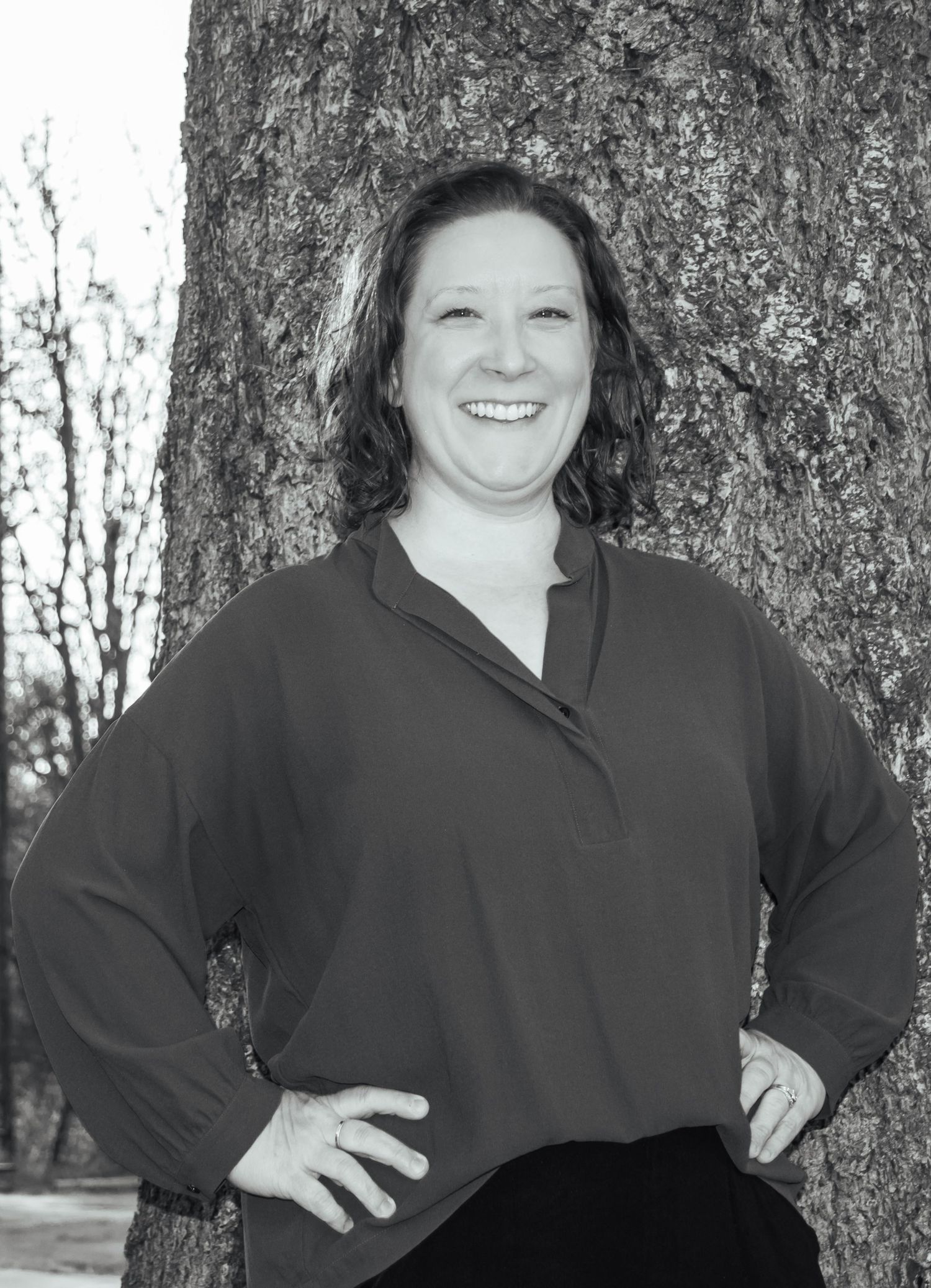 Black-and-white outdoor headshot of a smiling woman standing beside a tree.