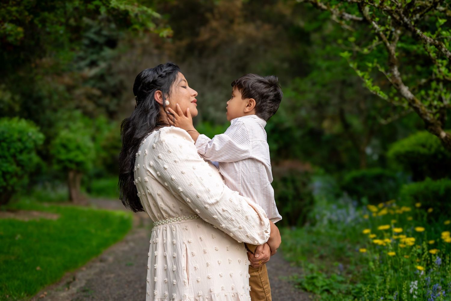 Mom and young son smiling at each other while cuddling on a garden path surrounded by lush greenery