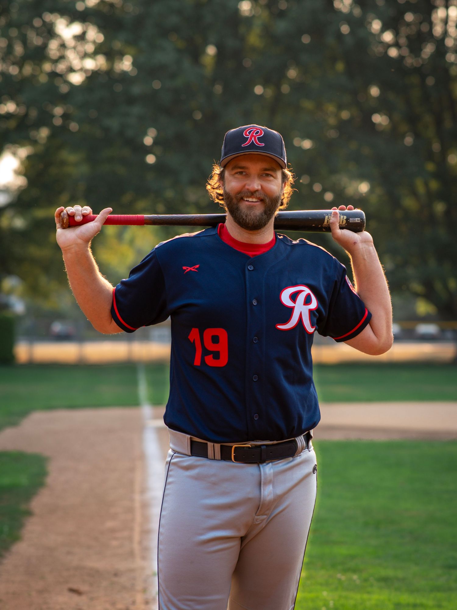 Baseball player smiling with a bat resting across his shoulders during an outdoor sports portrait session.