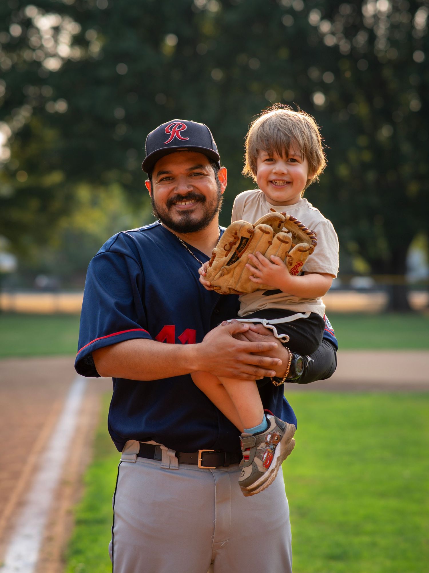 Baseball player holding a smiling young child with a baseball glove on the field at golden hour.
