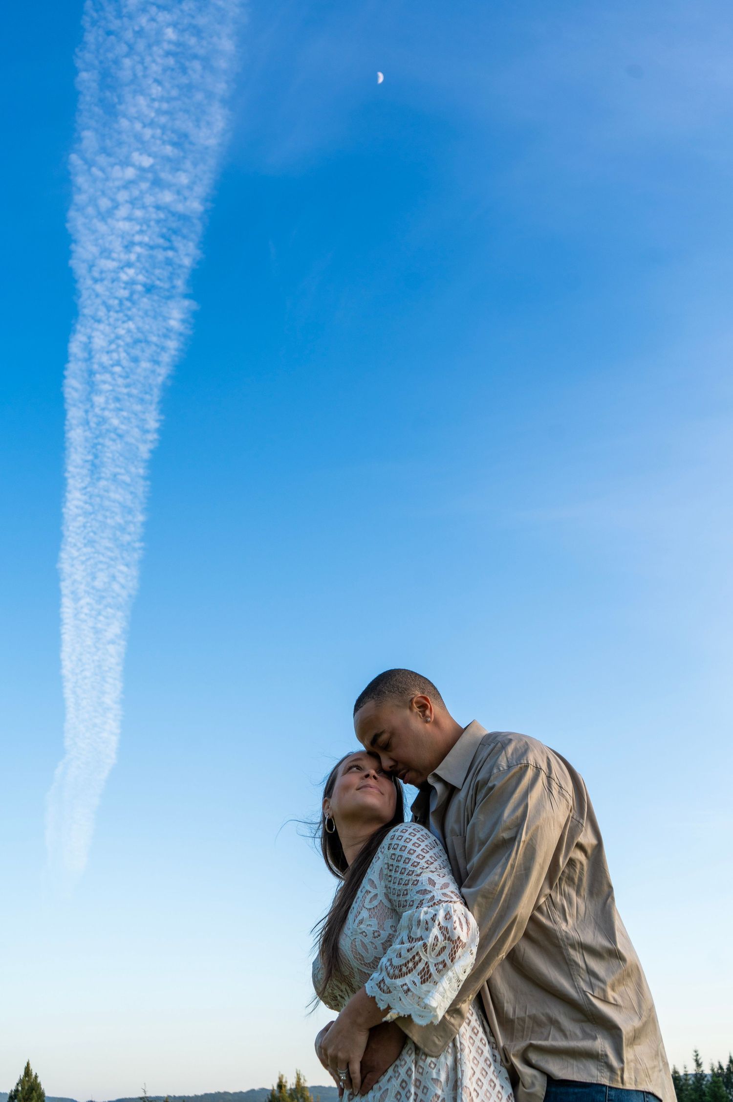 Couple cuddling beneath a blue sky and crescent moon during an outdoor couples photography session in Oregon