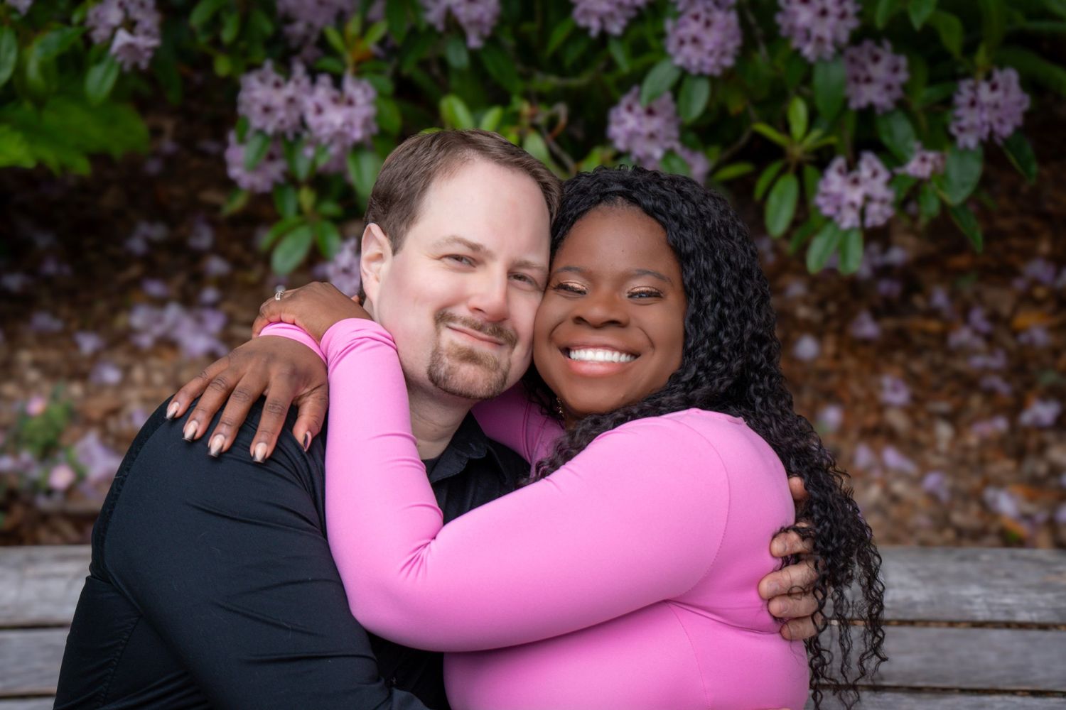 Couple smiling and embracing beneath blooming purple flowers during an outdoor couples photography session