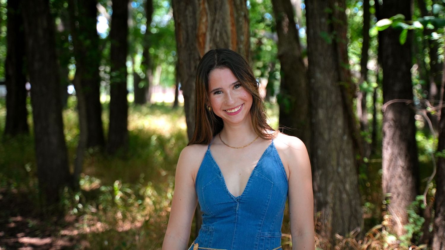 Young woman in blue dress smiling in a sunlit forest setting.