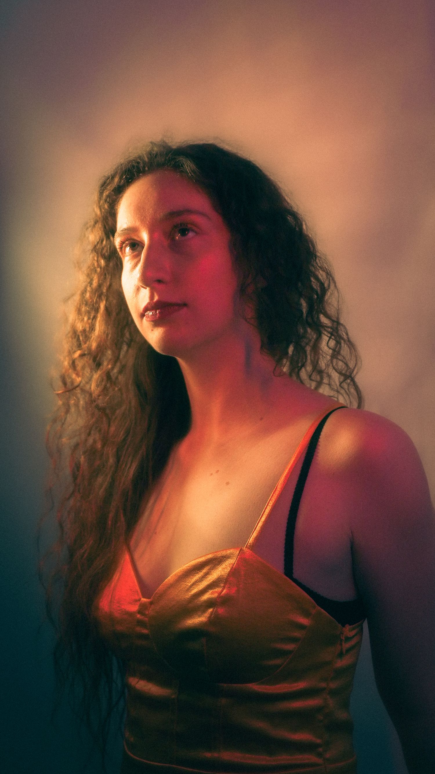 Young woman with curly hair poses in golden satin top under warm red and cool blue studio lighting.