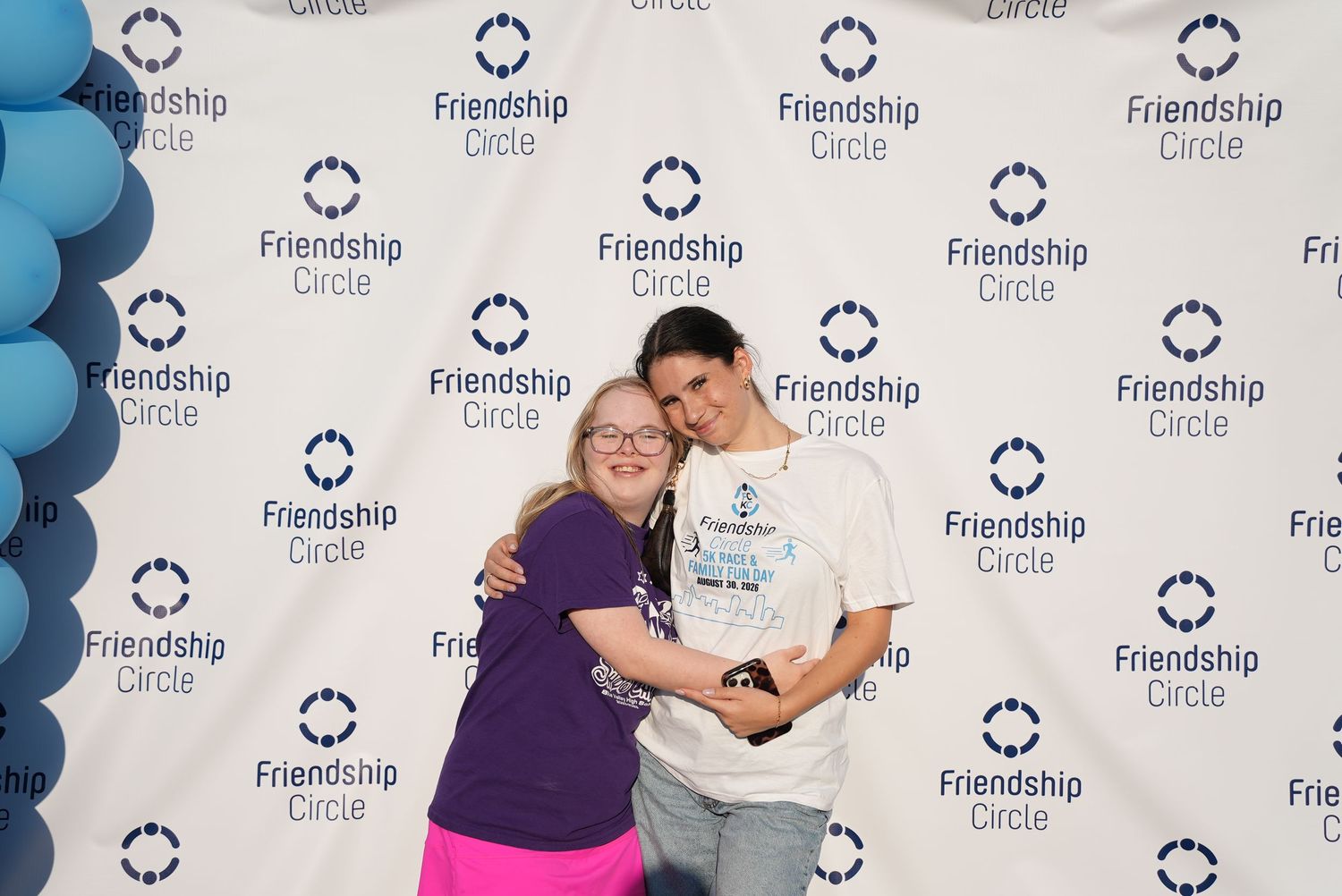 Two people pose together at a Friendship Circle event backdrop with blue balloons.