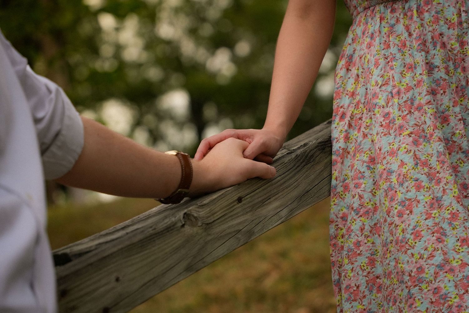 Couple holding hands over a rustic wooden fence, woman in floral dress, romantic outdoor setting.