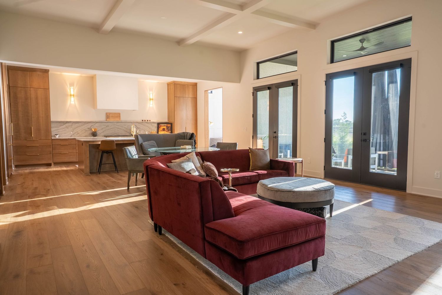 Modern open-concept living room with red velvet chairs, hardwood floors, coffered ceiling, and French doors leading to a balcony.