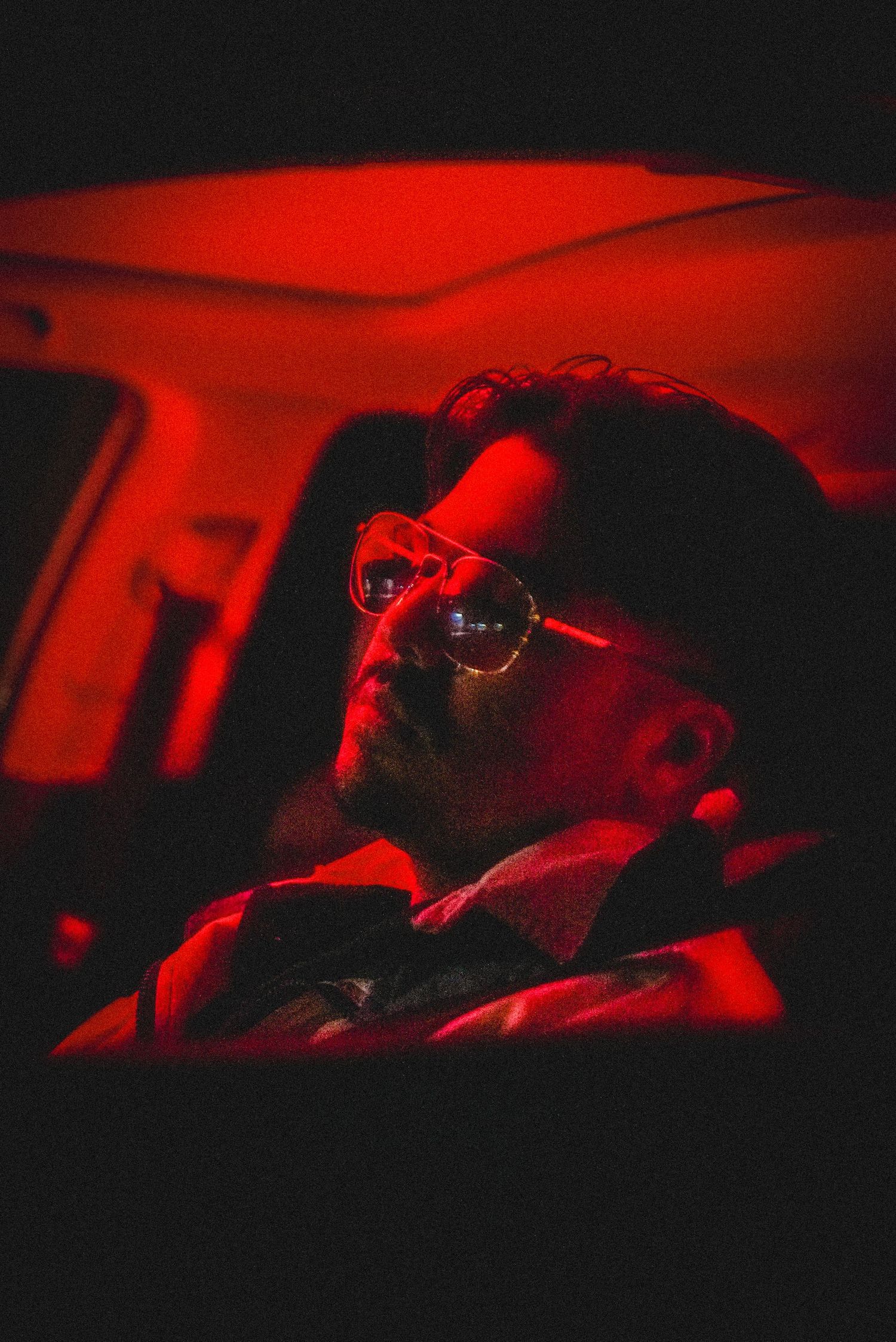 Man with beard and aviator glasses bathed in red light inside a dark car interior.