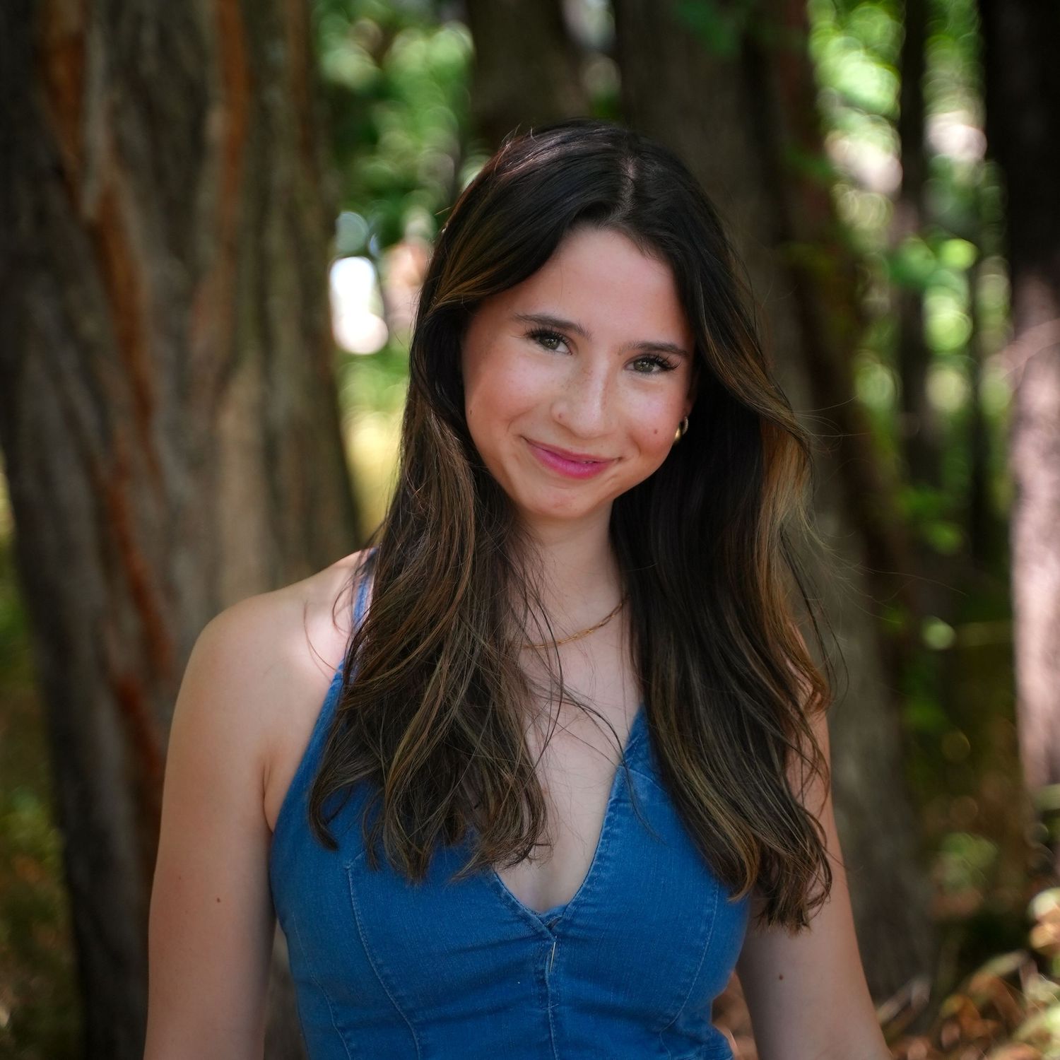 Young woman with long brown hair smiling in a blue top, posing outdoors among tall trees in a forest setting.