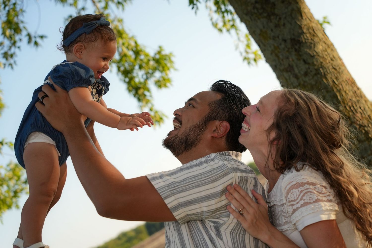 Happy family with parents lifting smiling baby outdoors under a tree on a sunny day.