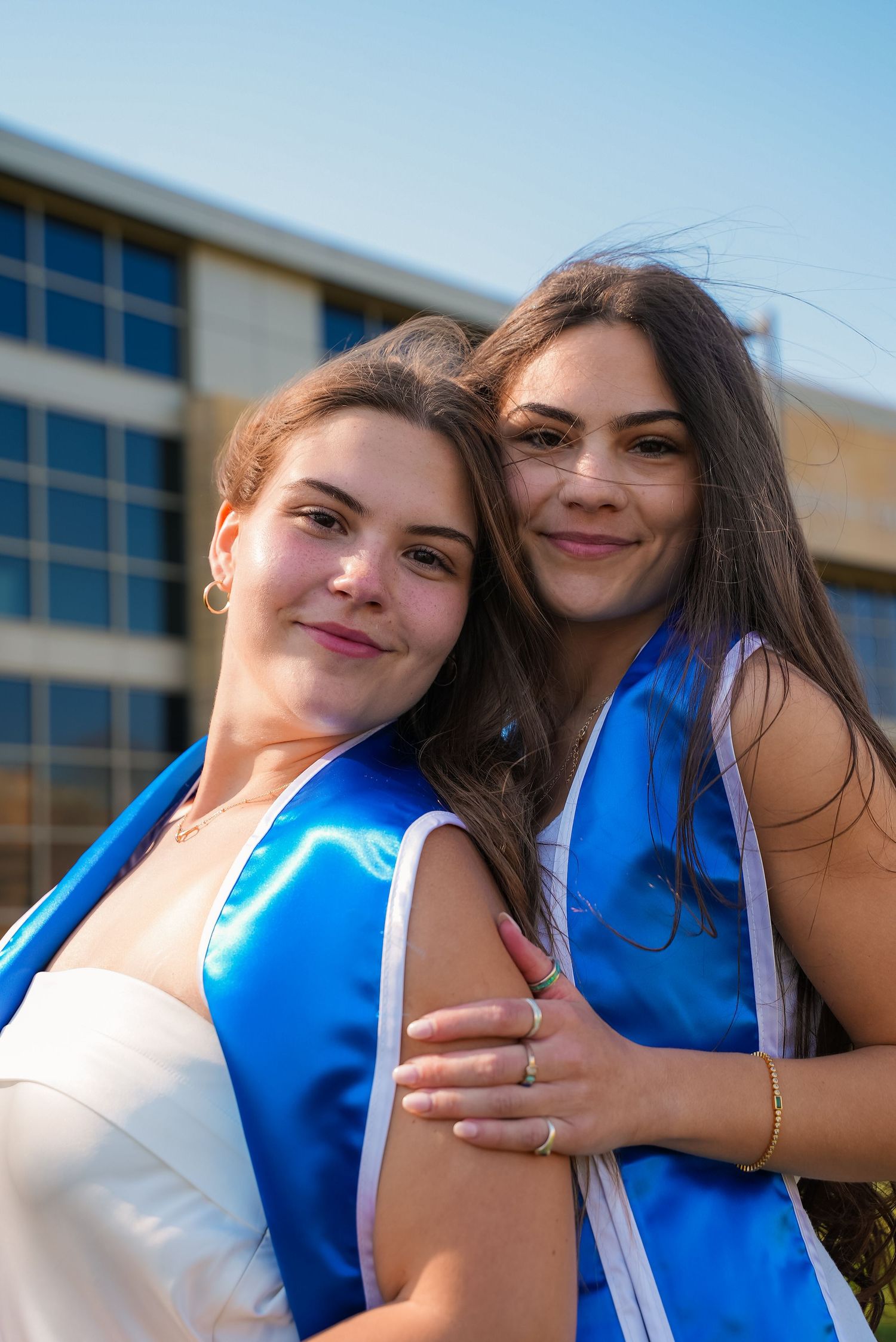 Two young women in blue graduation sashes smile and embrace outdoors on a sunny day.