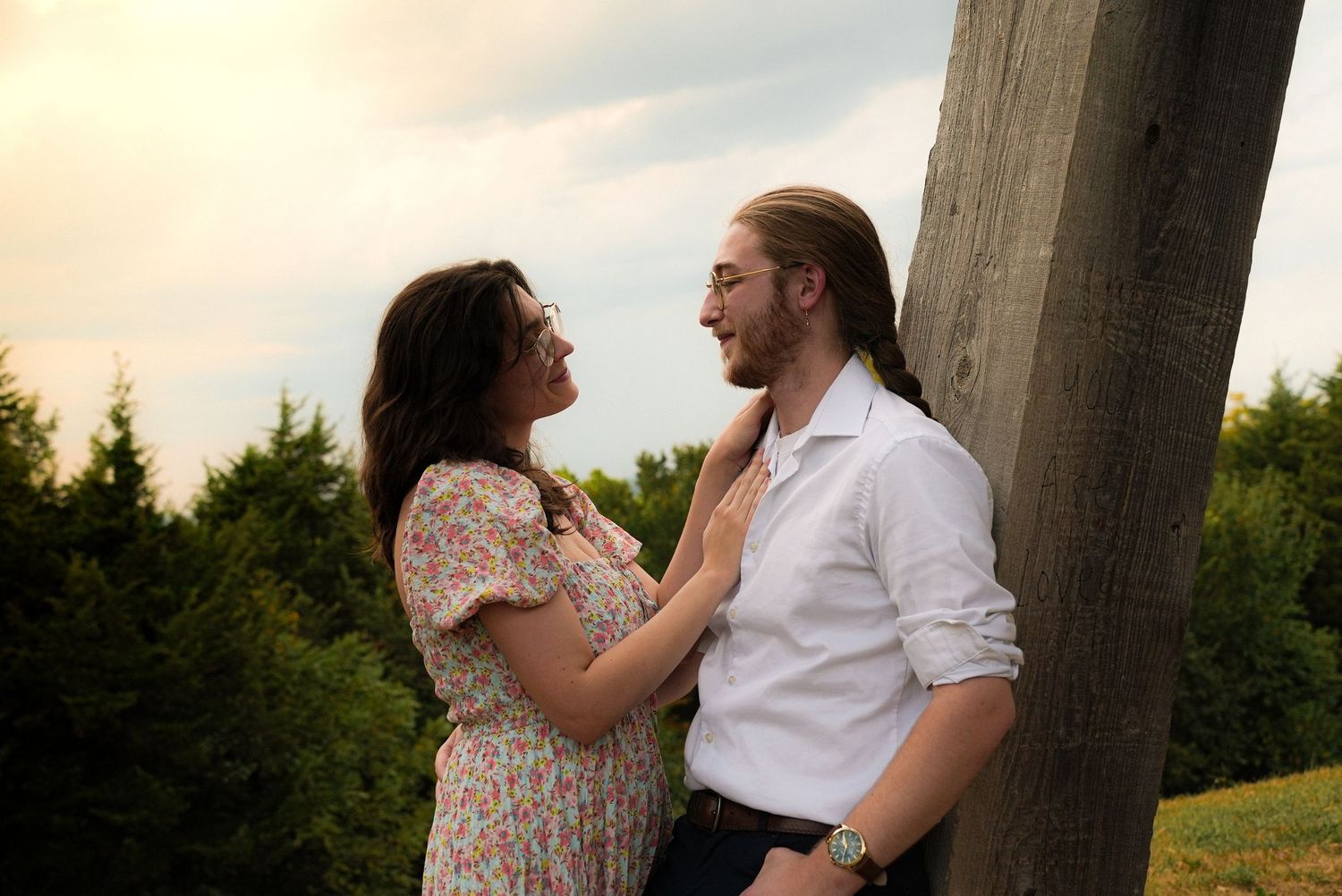 A couple shares a romantic moment outdoors, smiling at each other near a wooden post with green trees behind them.