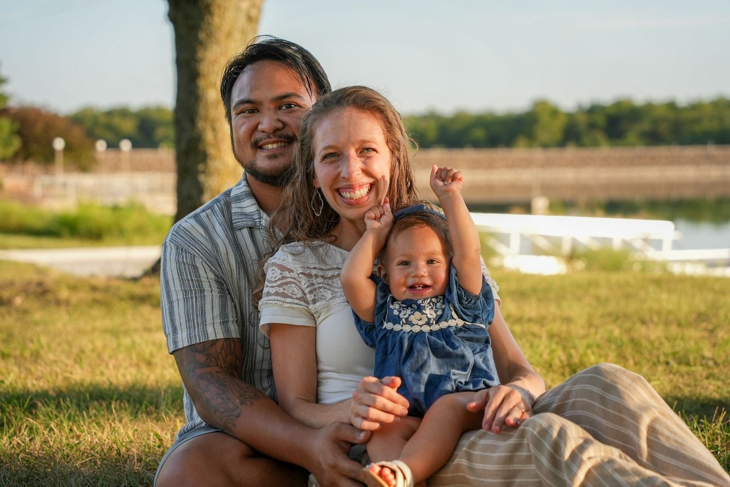 Happy family of three sitting on grass near a lake, parents smiling with their baby girl on a sunny day.