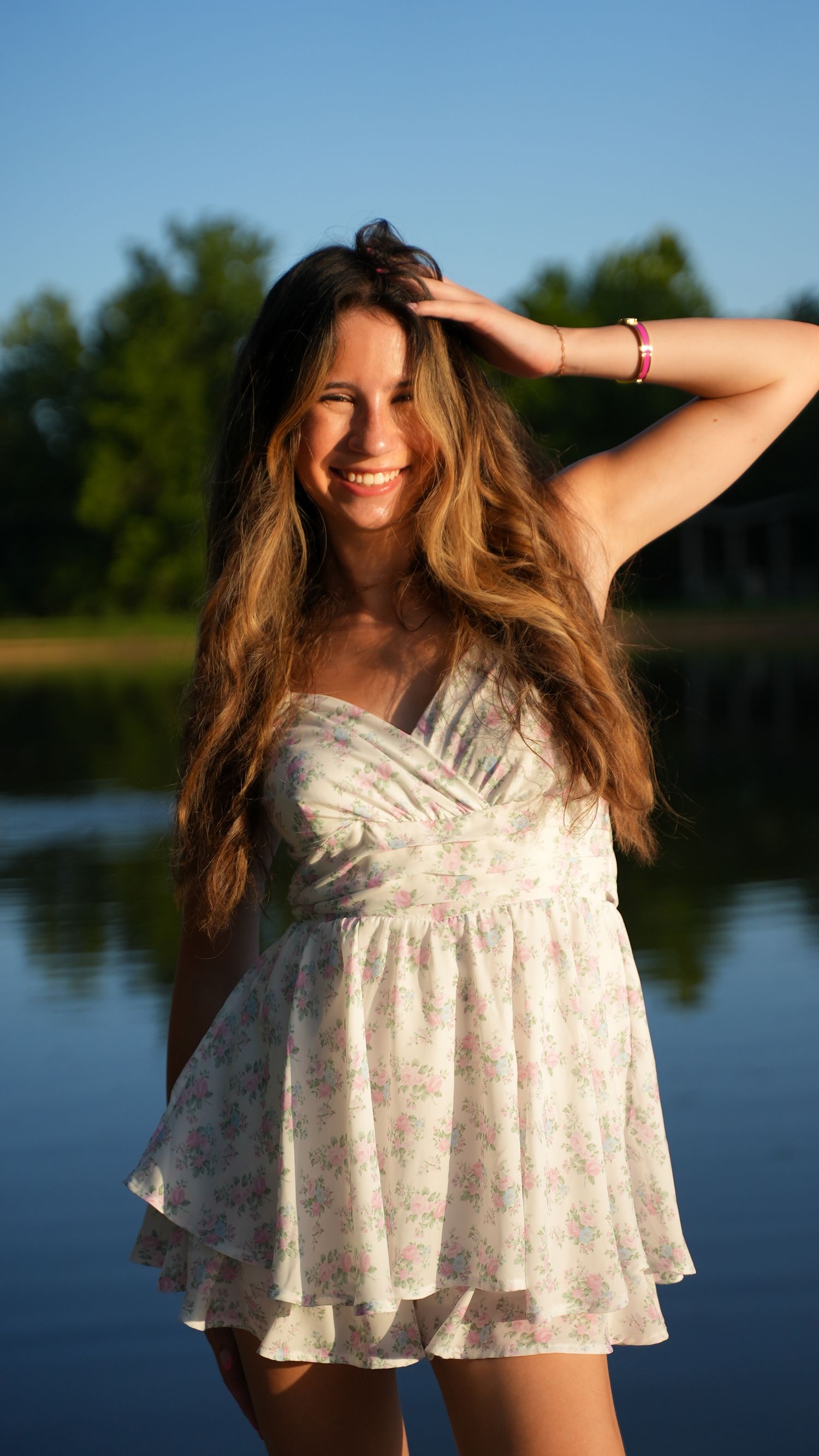 Smiling young woman in floral sundress poses by a lake at golden hour with trees in background.