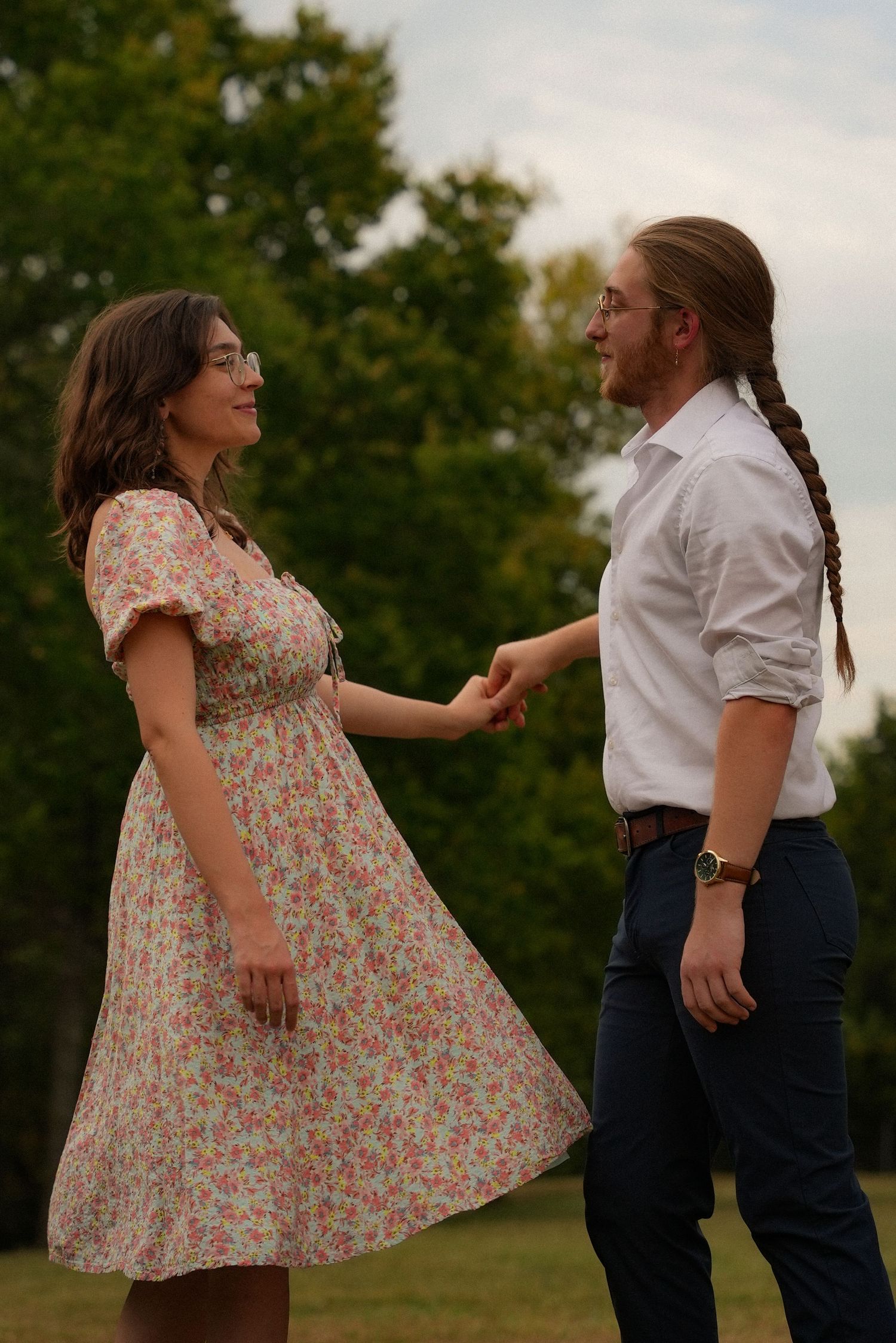 Couple holding hands in a park, woman in floral dress and man with braided hair in white shirt.