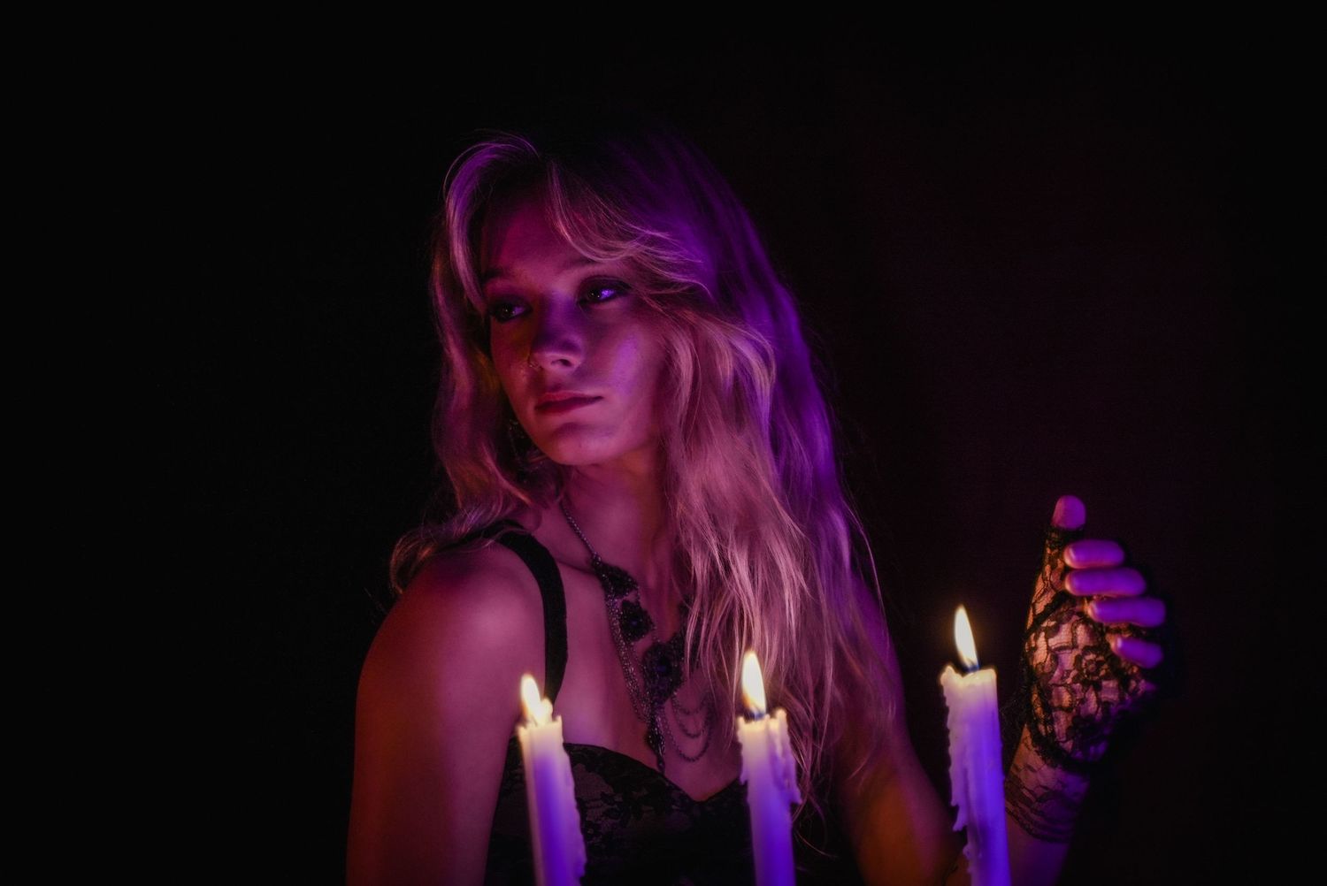 Blonde woman holding lit candles with purple lighting in a dark, mysterious Halloween witch-themed portrait.