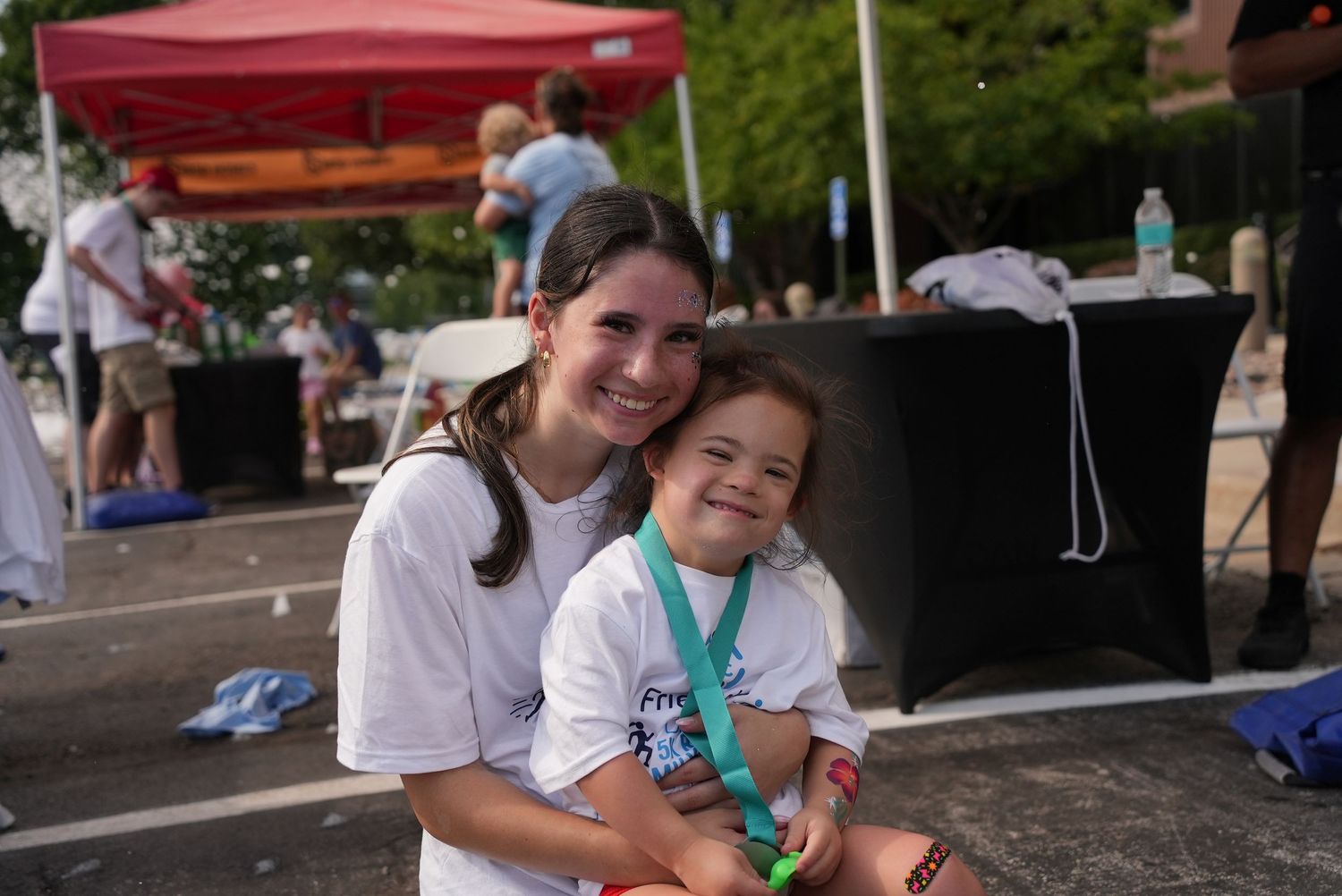 Two smiling girls in white t-shirts pose together at an outdoor charity walk event with a red tent in the background.