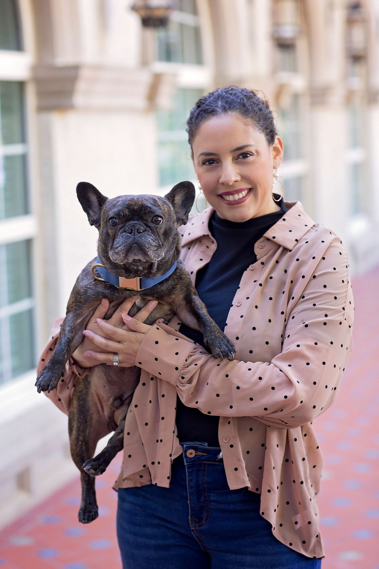 Smiling woman in polka dot jacket holds a dark brindle French Bulldog outdoors near a building.