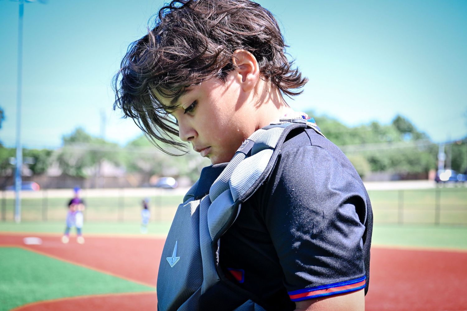Young baseball catcher in chest protector gear looks down thoughtfully during a youth baseball game.