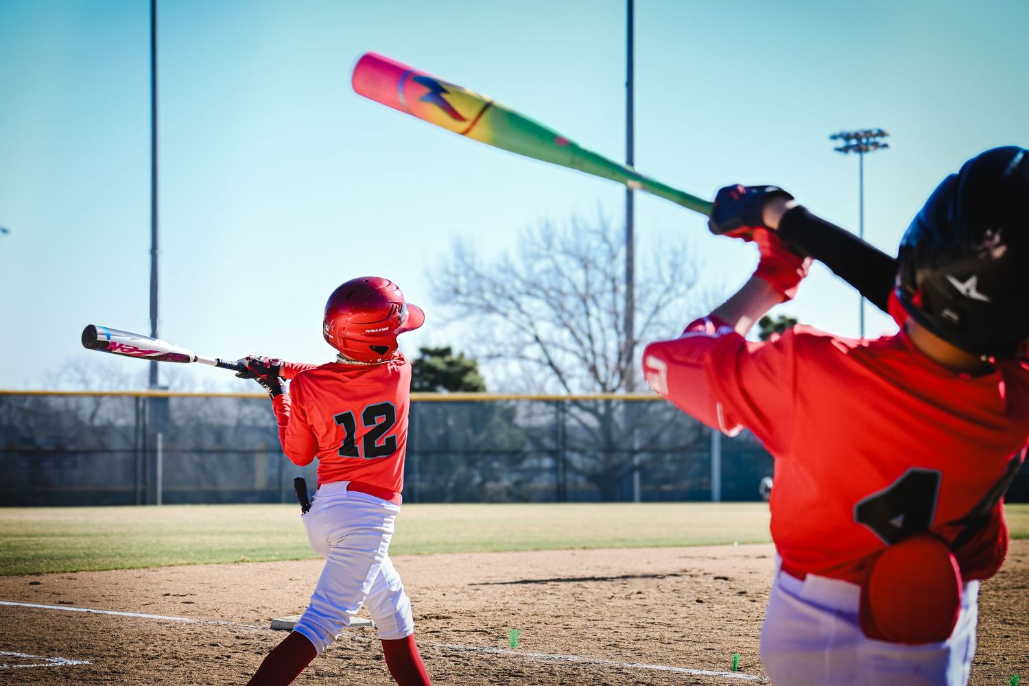 Two youth baseball players in red uniforms practice batting with colorful rainbow bat.