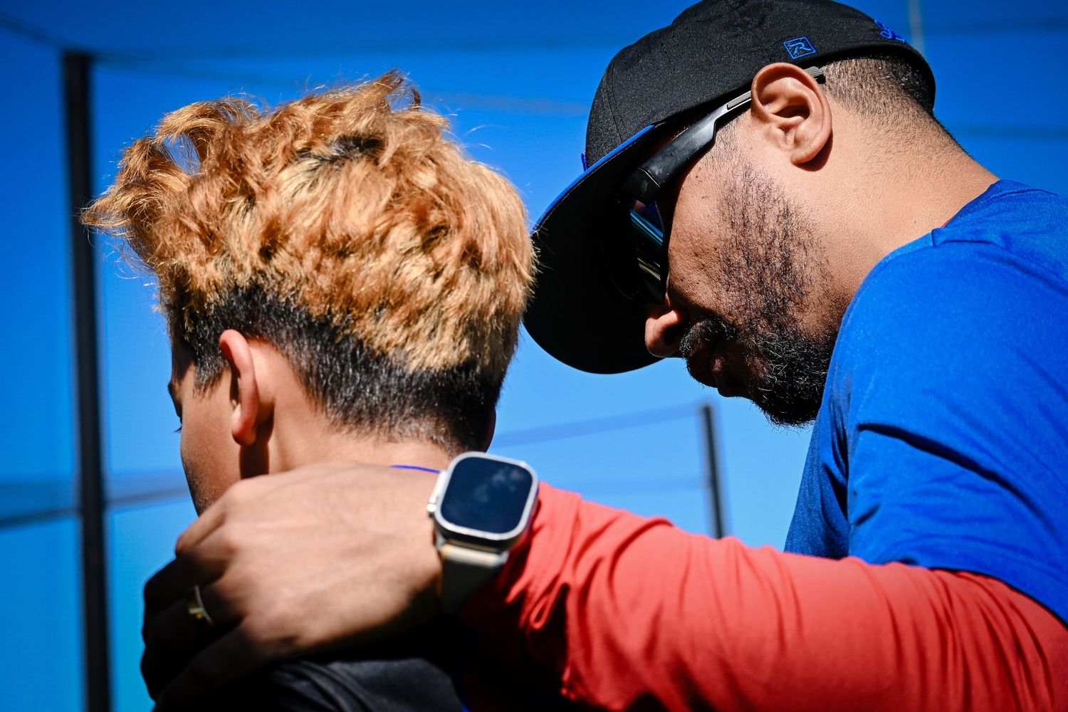 Coach in blue shirt and black cap gives close guidance to a young baseball player outdoors.