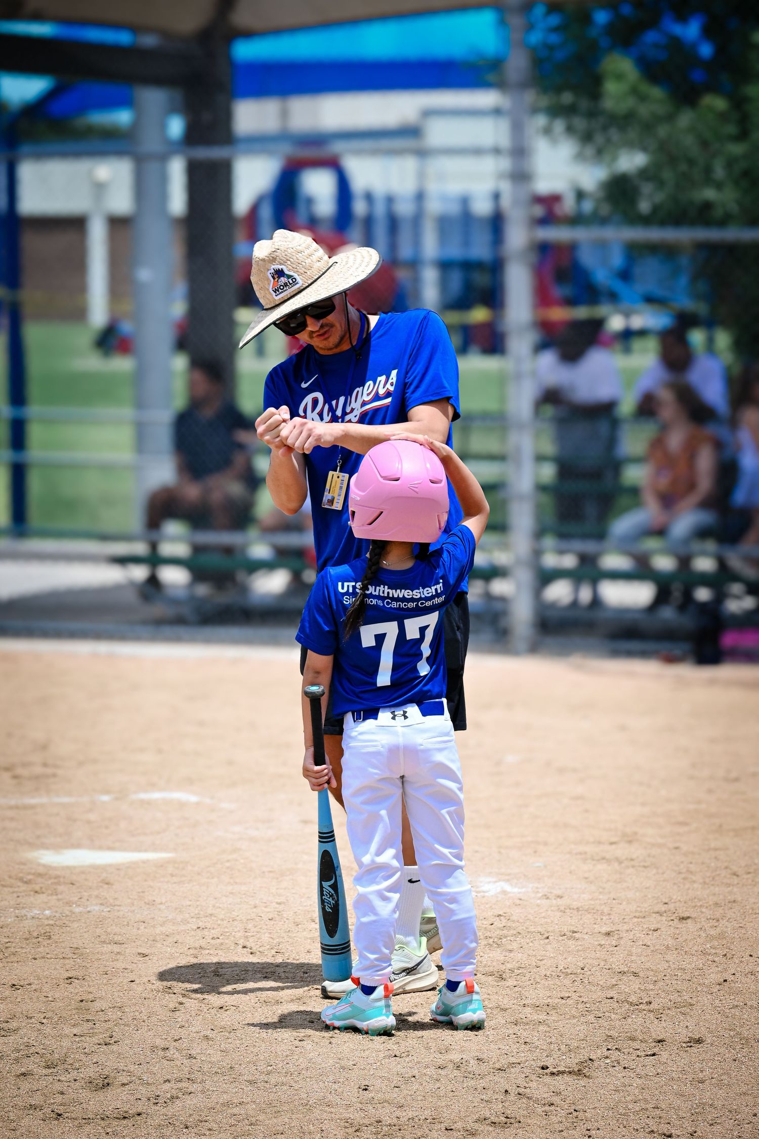 Baseball coach in straw hat helps young player #77 with batting stance during youth baseball practice.
