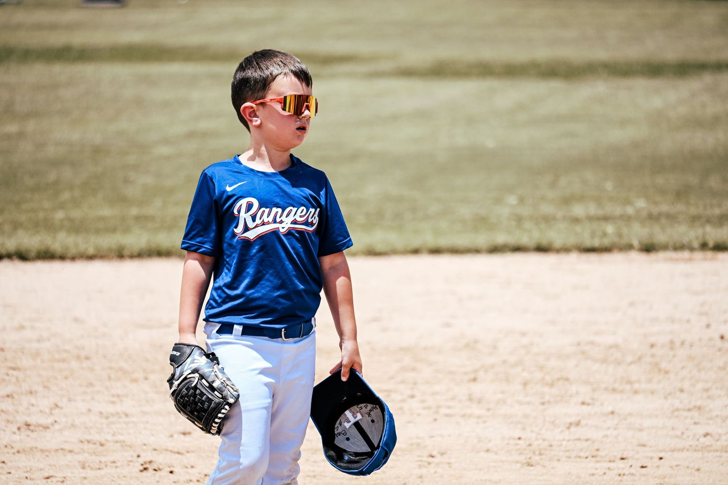 Young Rangers baseball player in blue uniform holds glove and helmet on sunny baseball diamond.