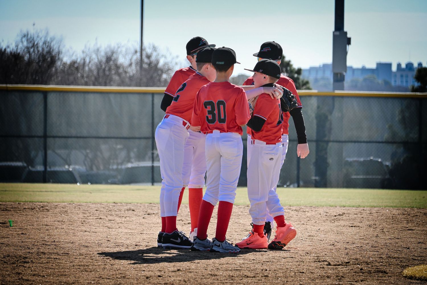Youth baseball players in red and white uniforms huddle together on the pitcher's mound during a game.