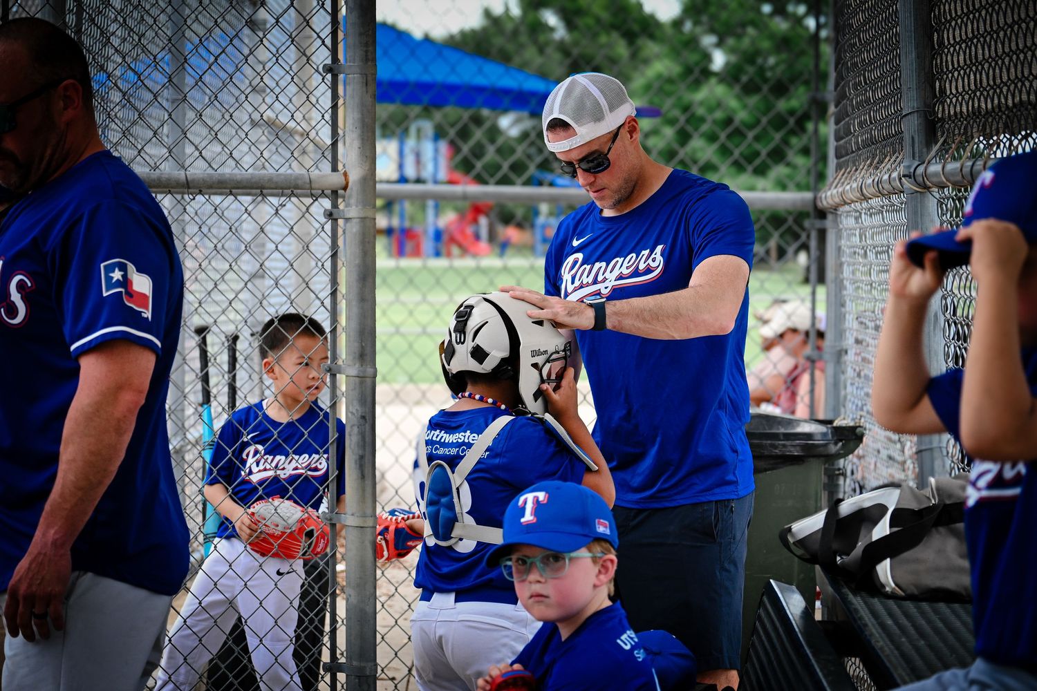 Little league baseball coach helping young catcher put on gear in dugout during Rangers game.