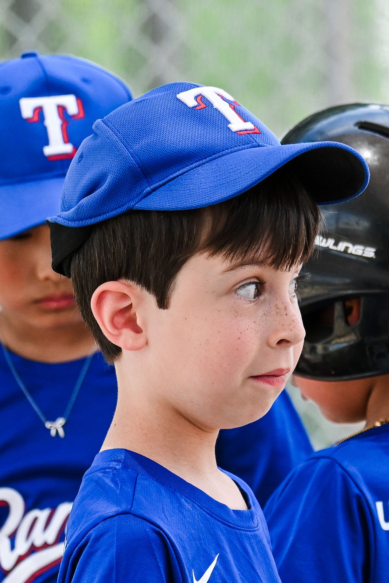 Young boy in blue Texas Rangers baseball uniform and cap looks up attentively during a youth game.