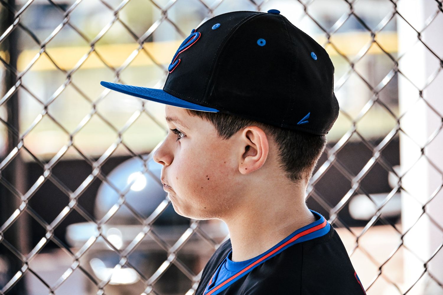 Young boy in black and blue baseball cap gazes through a chain-link fence at a baseball game.