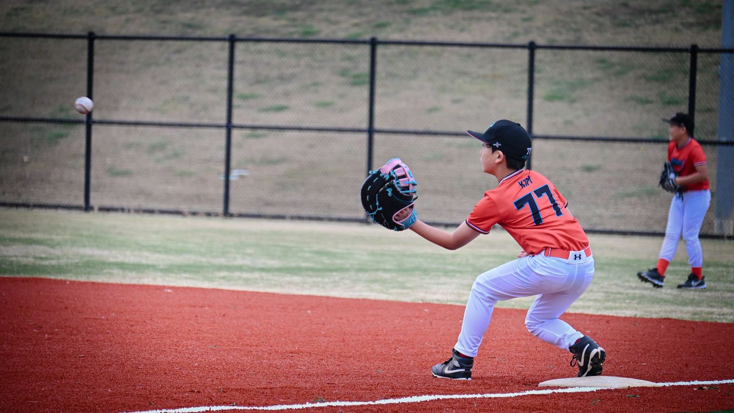 Youth baseball player in orange number 74 jersey catching a ball on a red turf infield during a game.