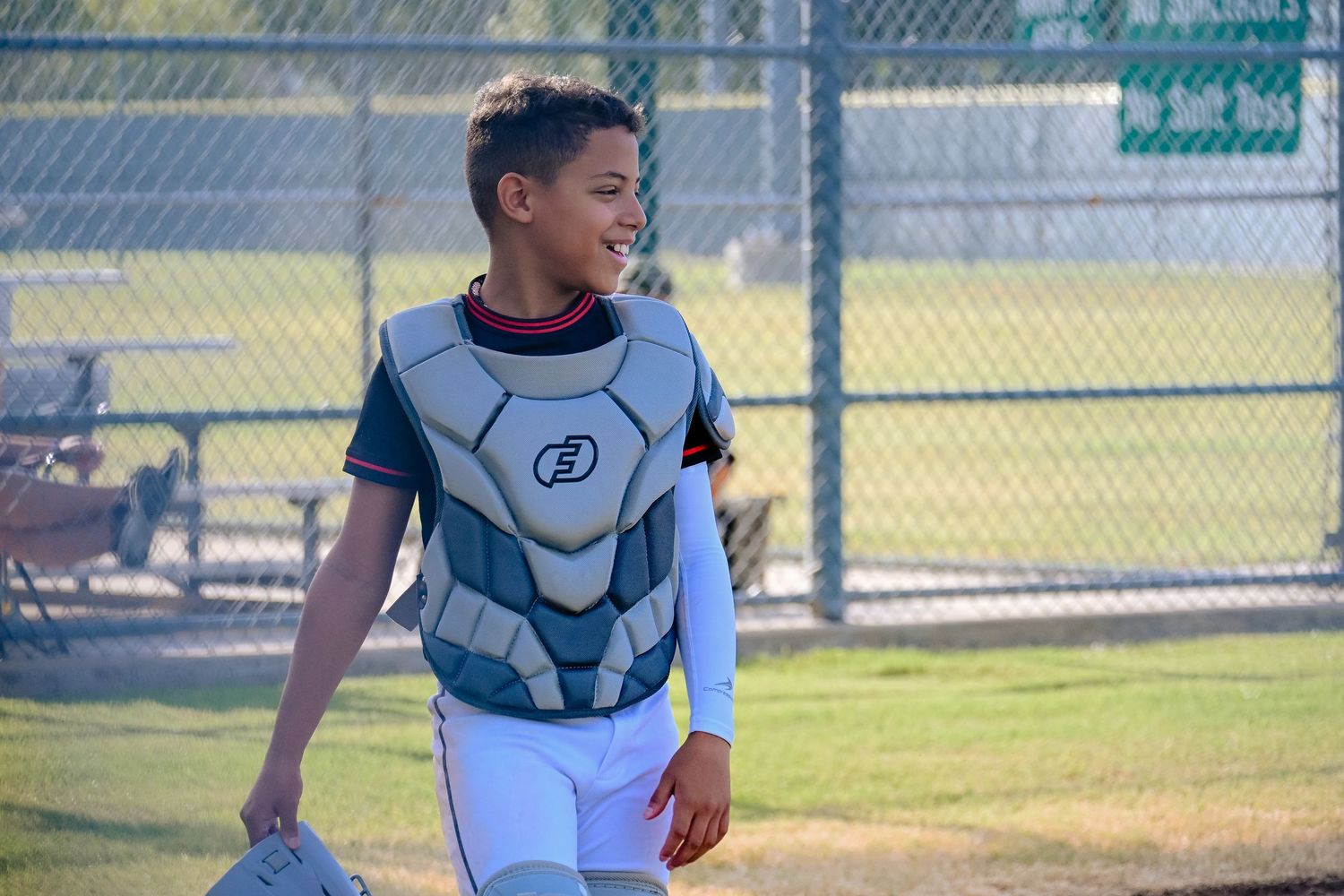 Young baseball catcher in chest protector gear smiling on sunny field.