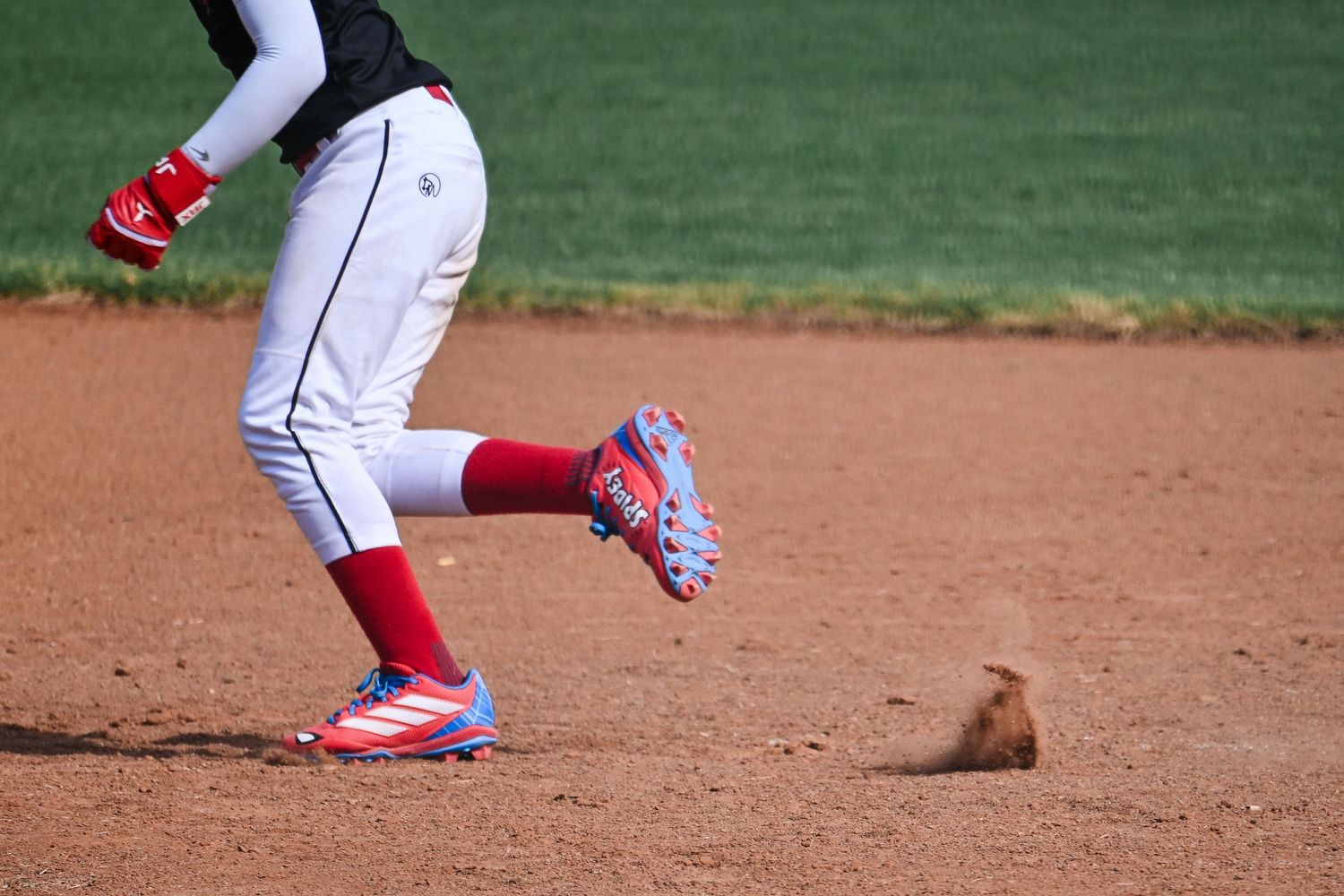Youth baseball player in white uniform with red socks running past base with small animal nearby.