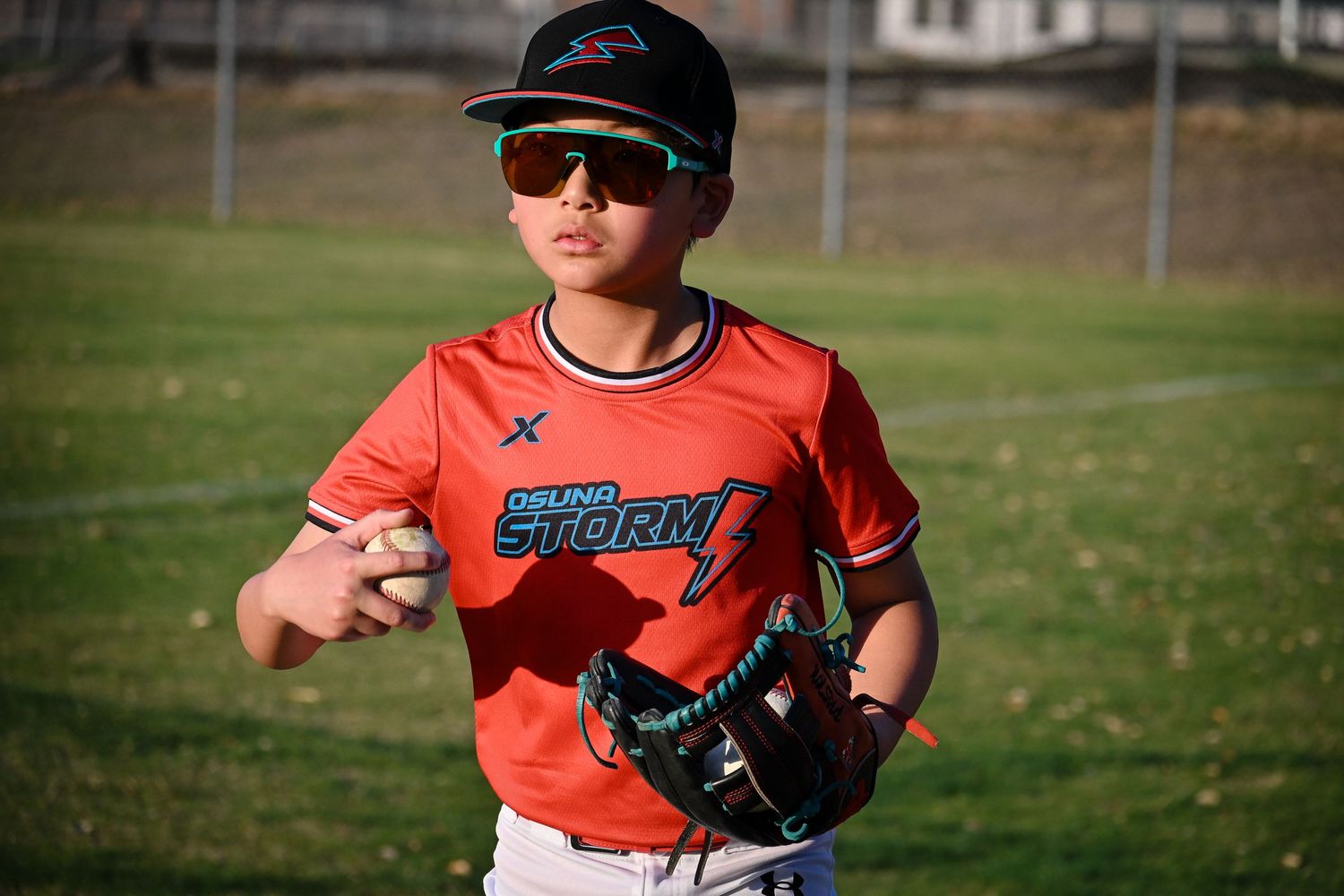 Youth baseball player in orange Inland Storm jersey holding a ball and glove during practice outdoors.