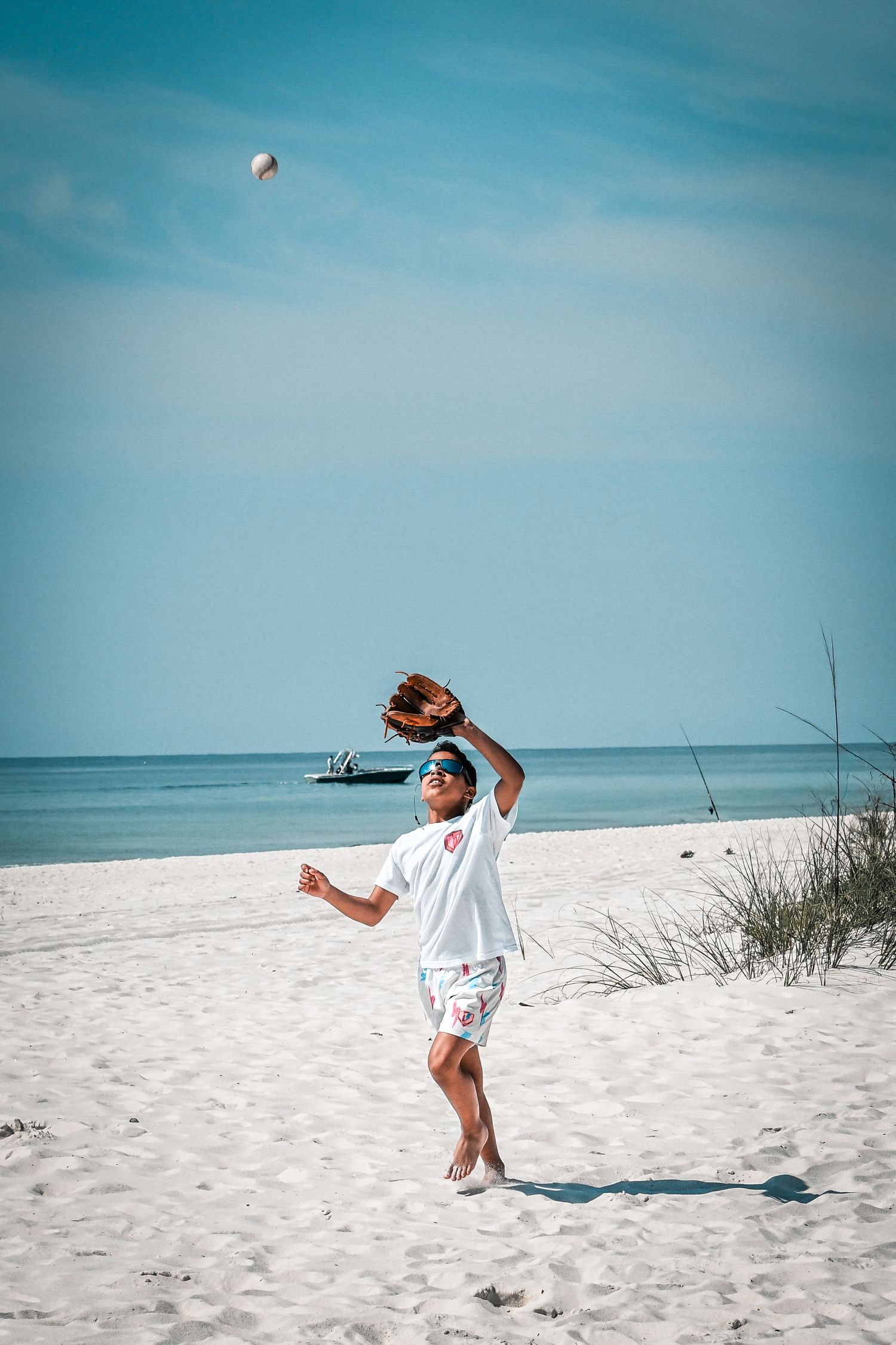 Young man in white shirt catches a baseball on a sunny beach with blue ocean in background.