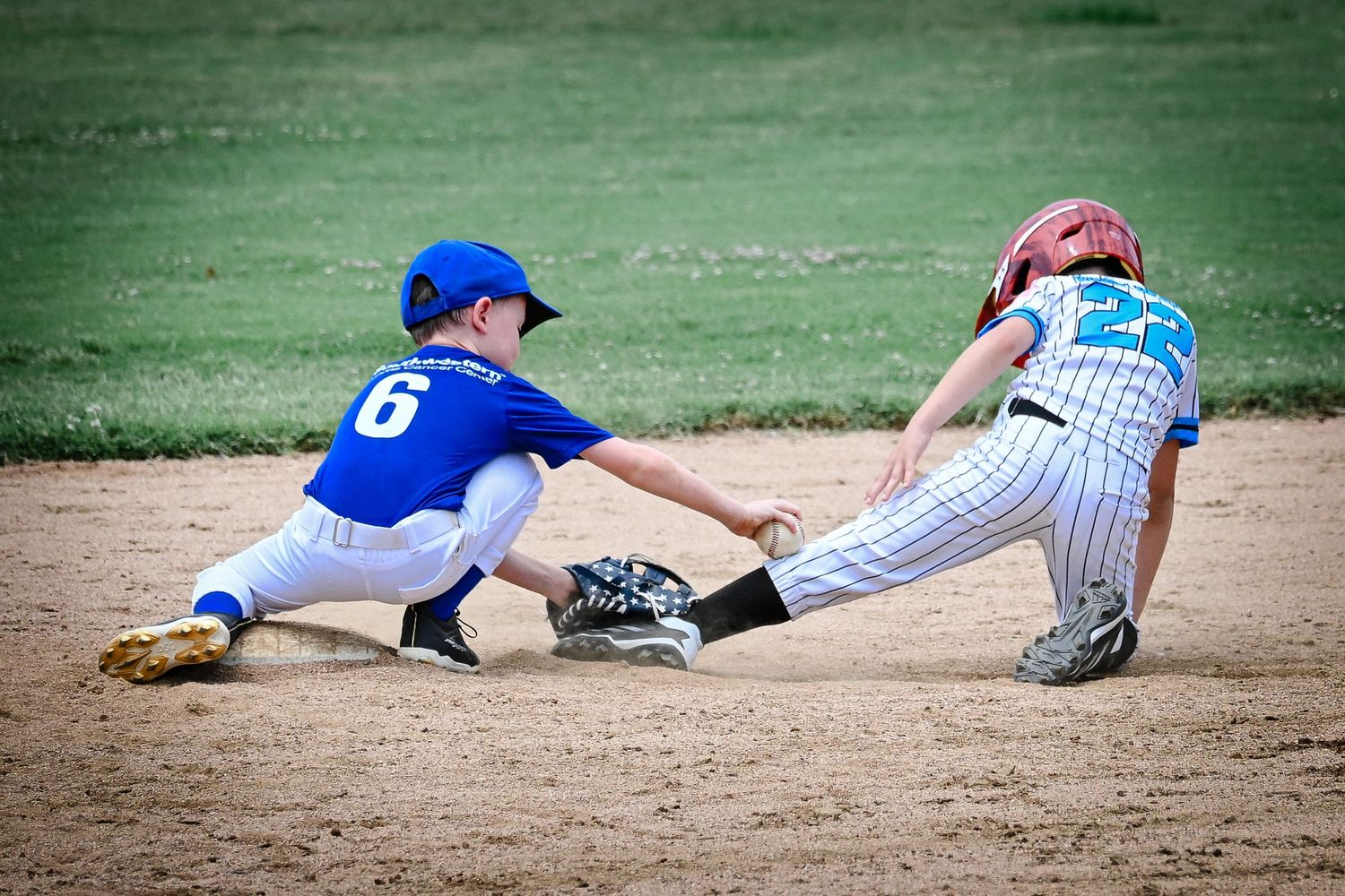 Youth baseball player #6 in blue stretches to tag out sliding runner at second base.