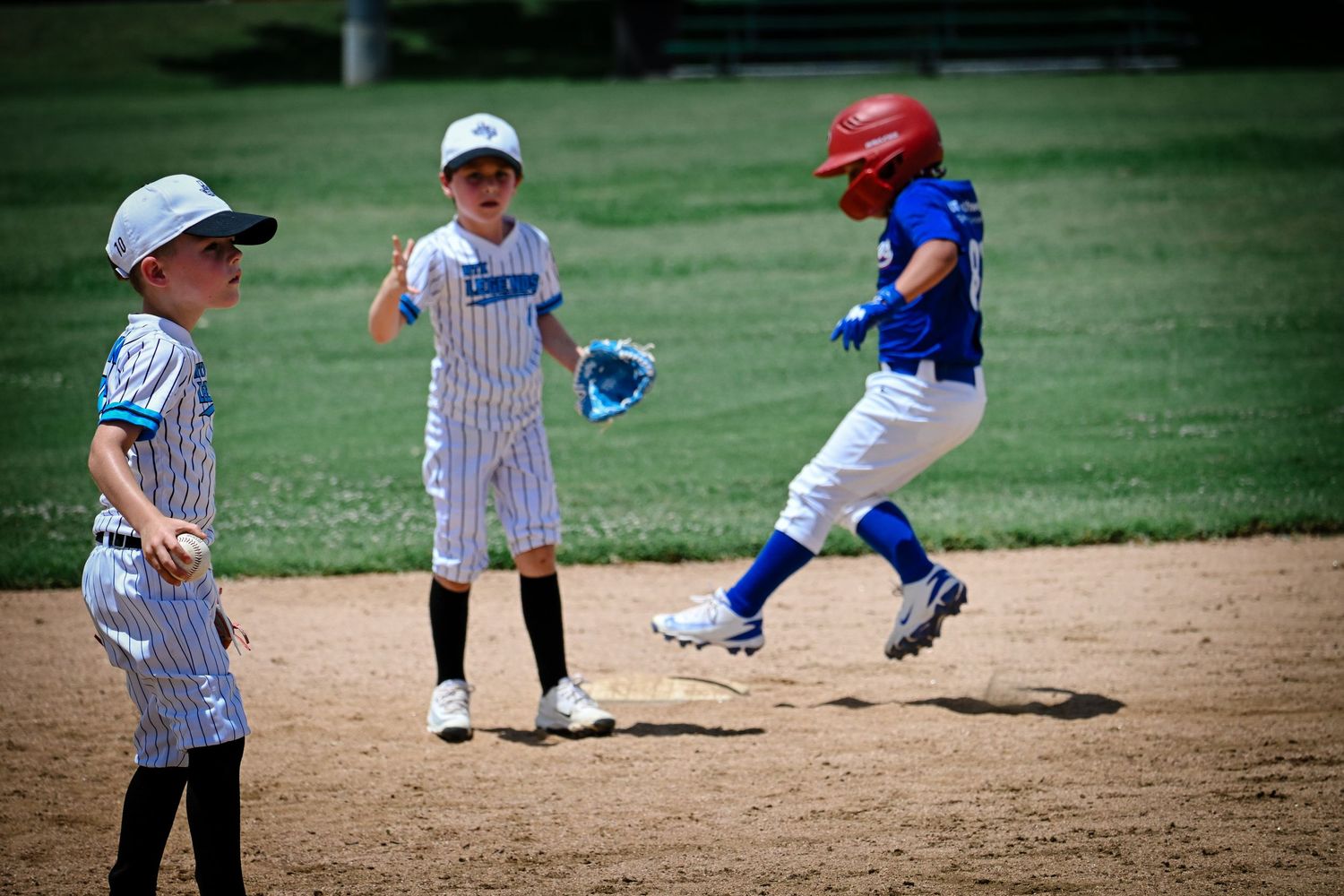 Young baseball player in blue Rangers uniform running bases past two opposing team players.