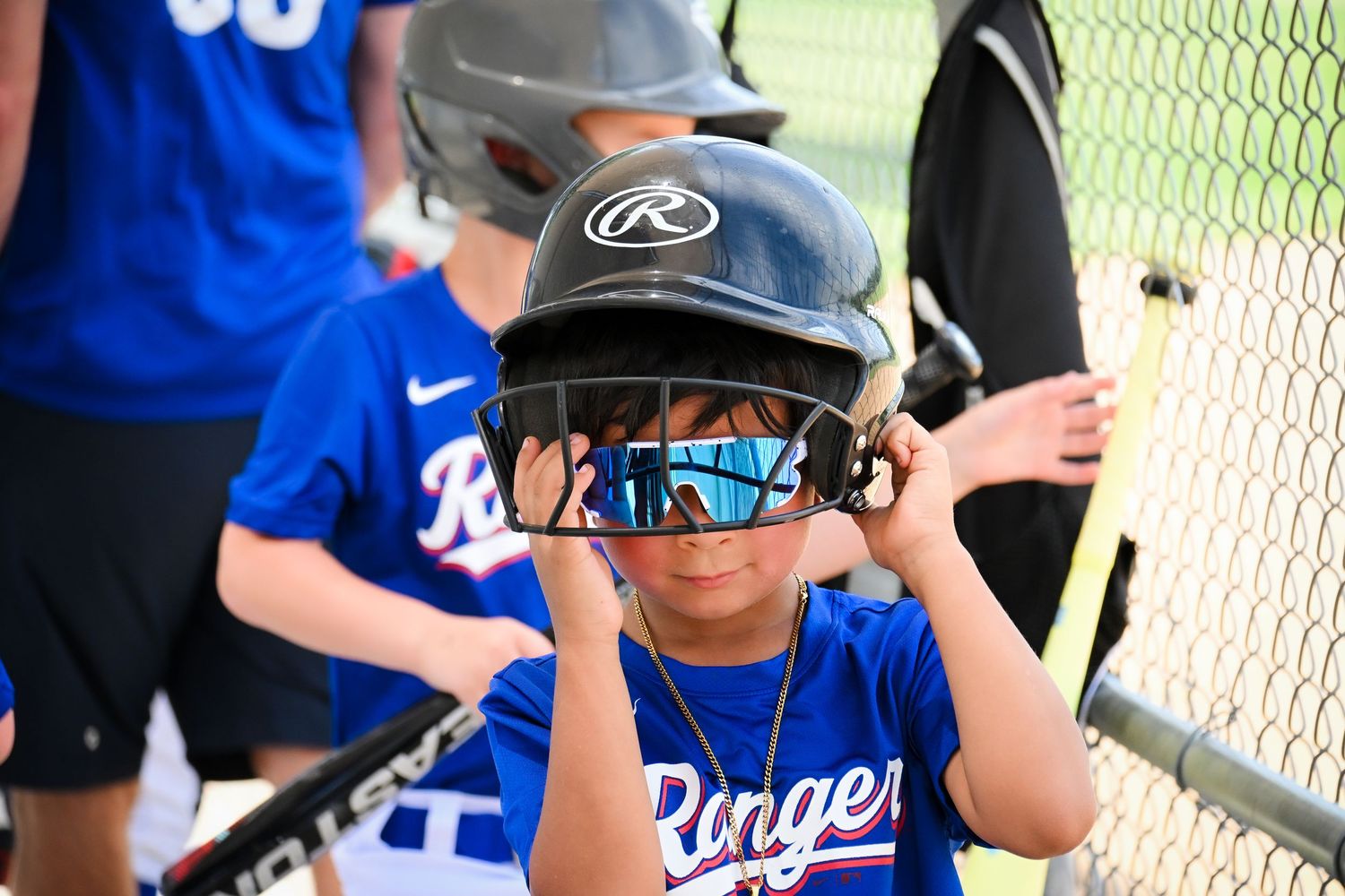 Young baseball player in Rangers uniform adjusting black Rawlings batting helmet with sunglasses on sunny game day.