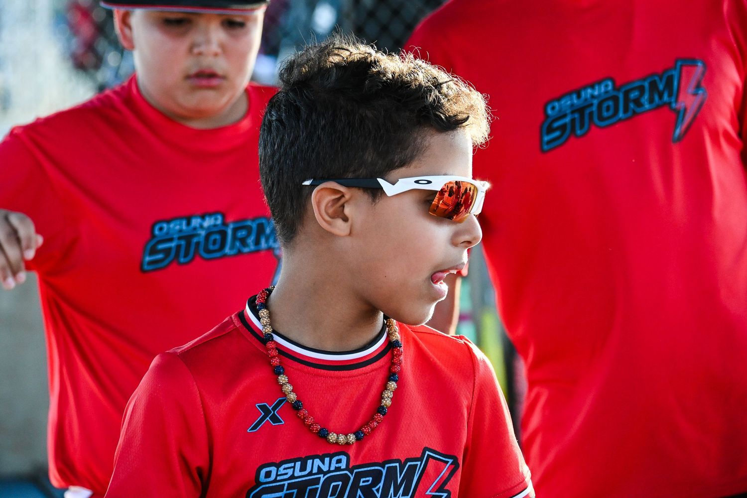 Young baseball player wearing Osuna Storm red jersey and sunglasses stands focused during a youth game.