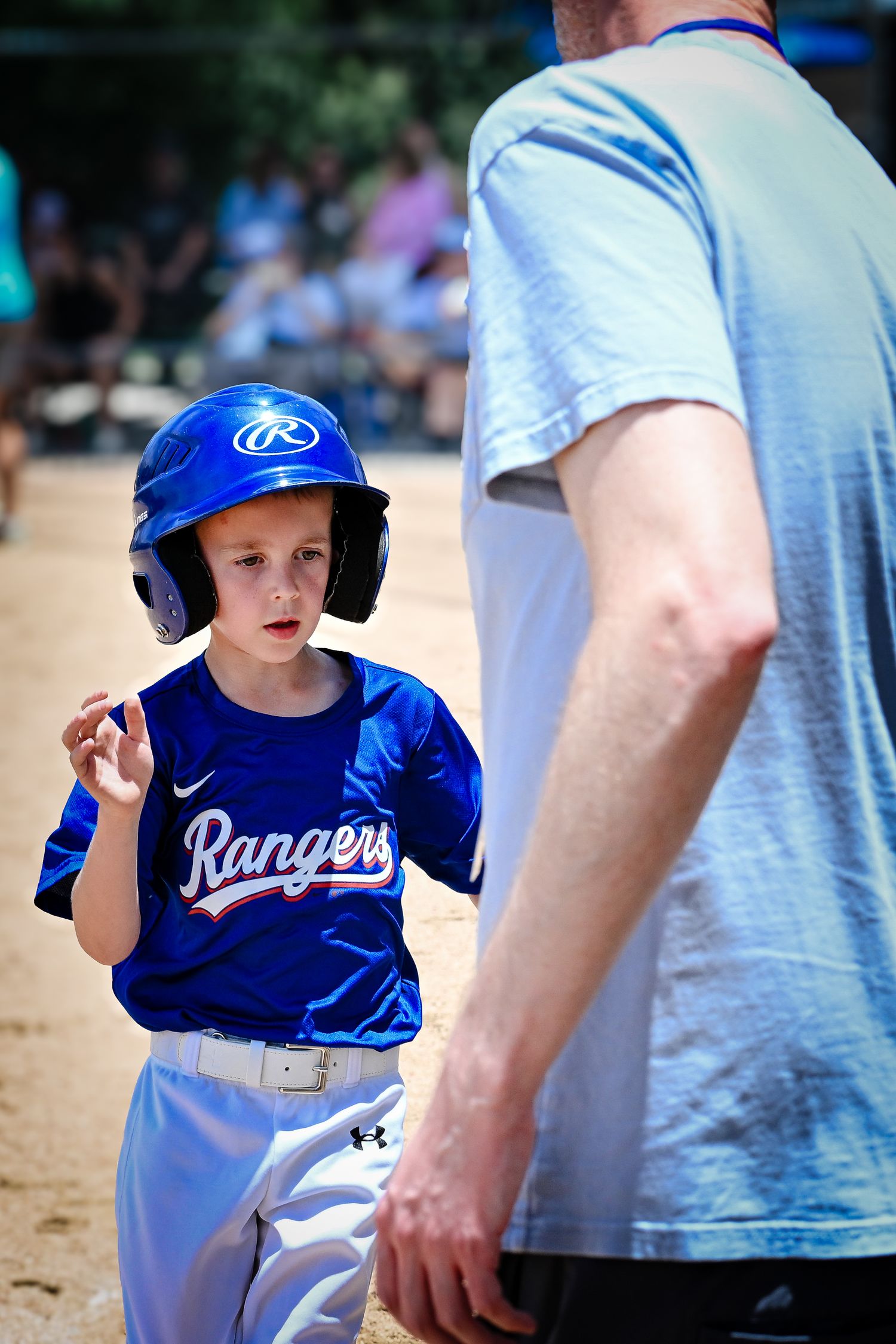 Young boy in blue Rangers baseball helmet and jersey listening to coach during little league game.