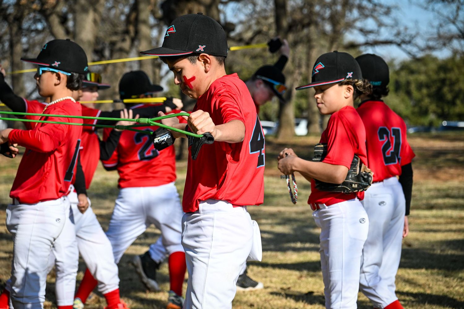 Youth baseball players in red uniforms stretch with resistance bands during warm-up drills on a sunny field.