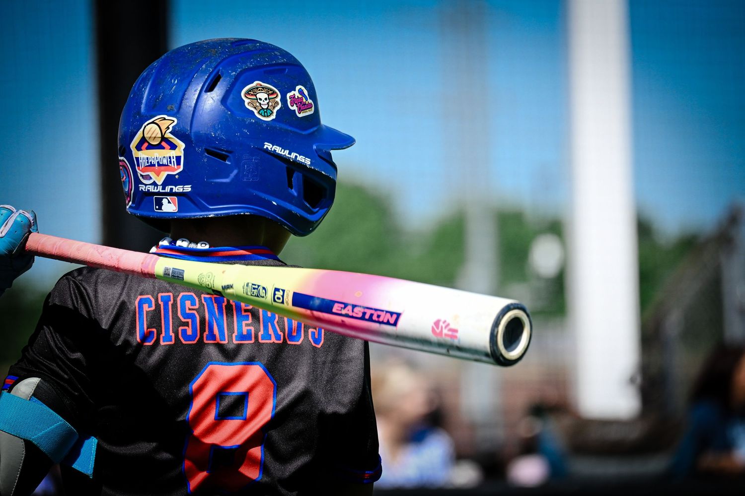 Young baseball player in blue helmet holds an Easton bat, wearing a Cisneros jersey during a youth game.