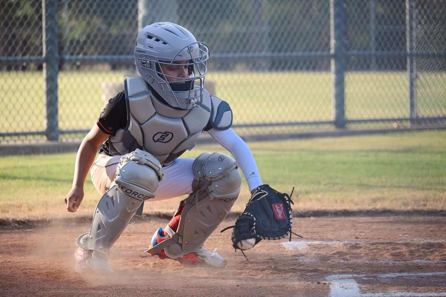 Youth baseball catcher in full gear crouching at home plate ready to catch pitch in evening game.