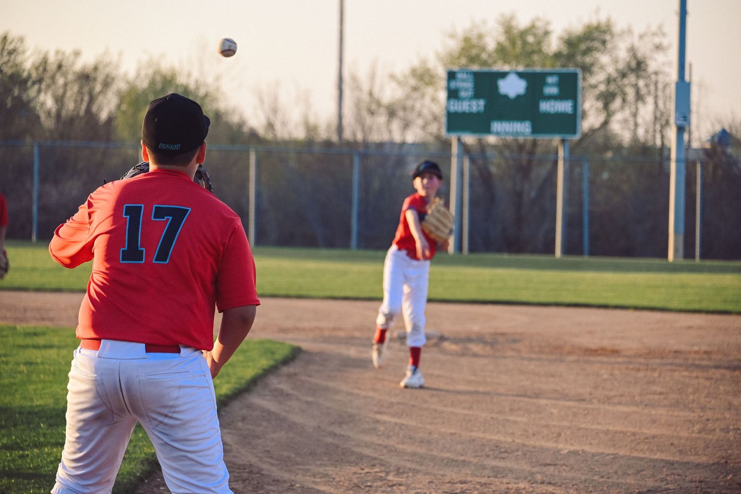 Two youth baseball players in red uniforms practice throwing on a baseball field at sunset.