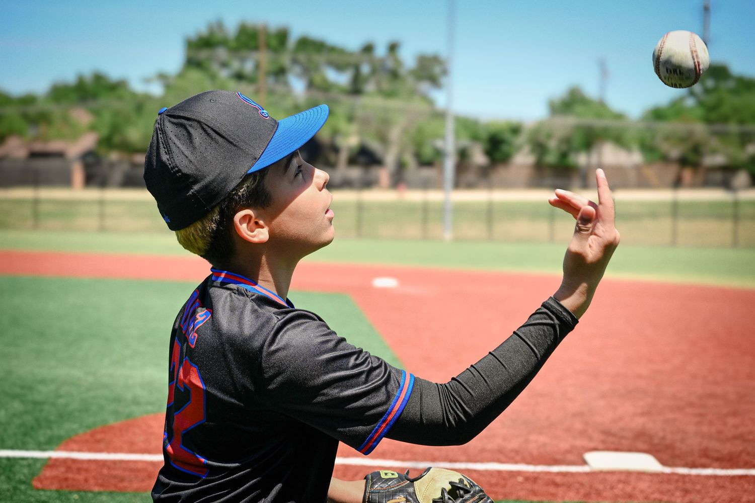 Young baseball player in blue cap tossing ball skyward on sunny outdoor baseball field.
