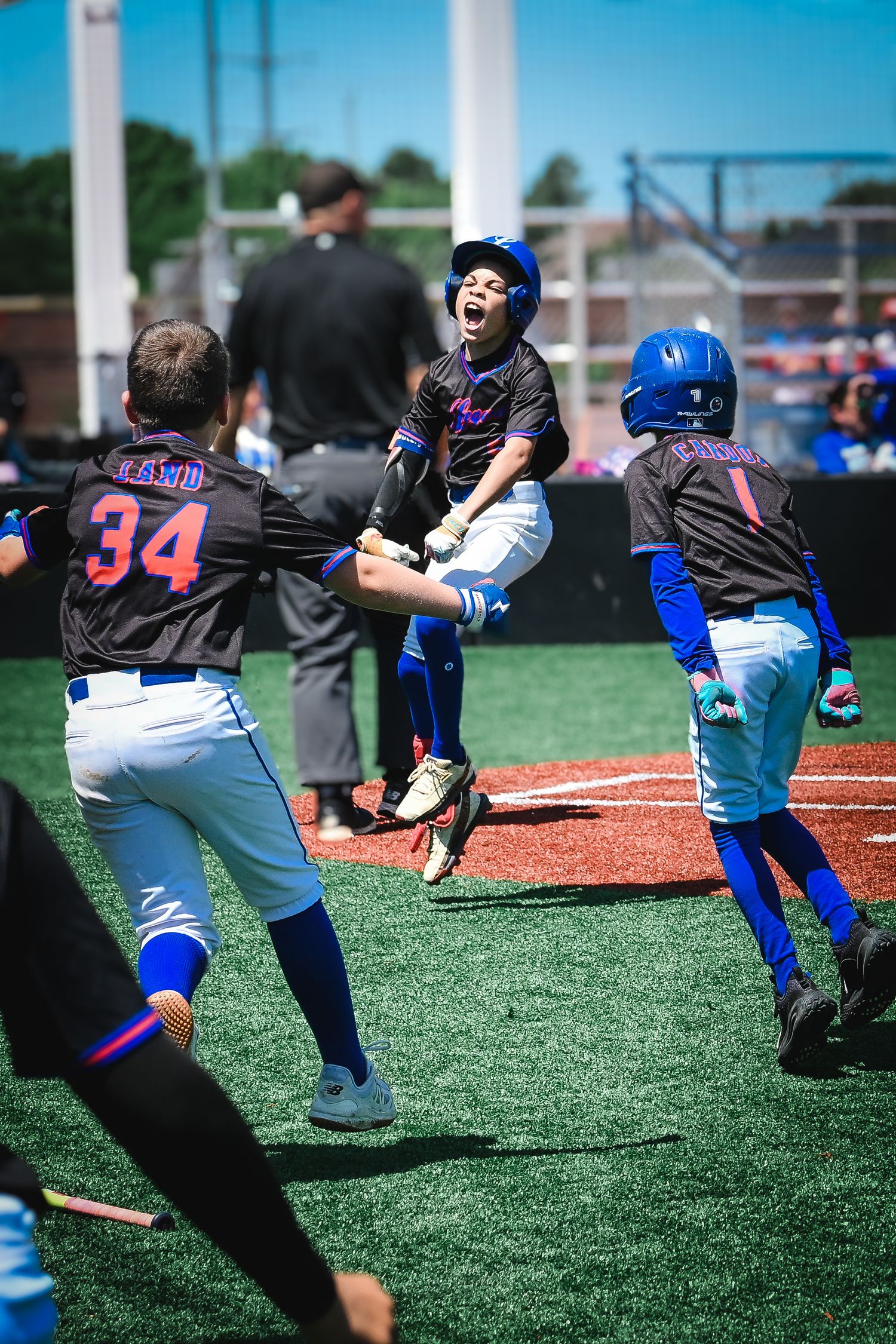 Excited young baseball player jumps celebrating near home plate with teammates cheering around him.